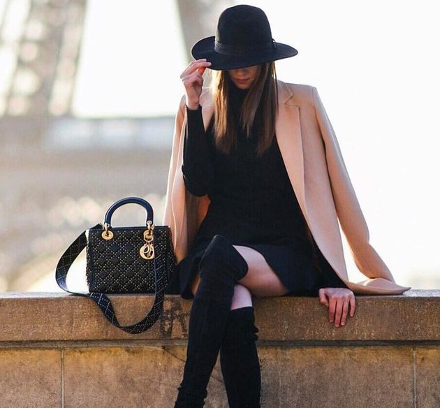 A woman in a black hat, black dress, and beige coat sitting with a designer handbag.